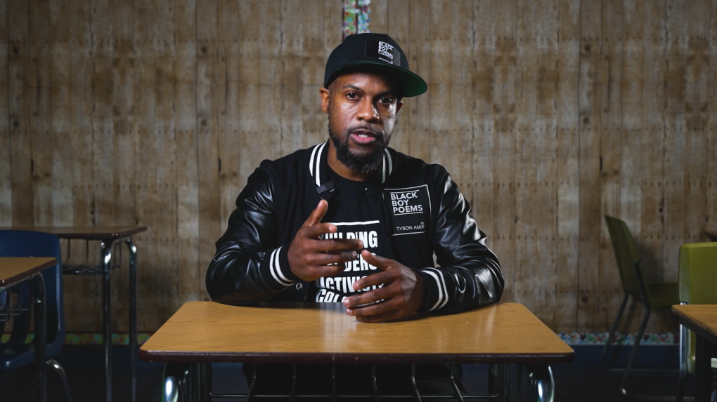 An African-American man in a black hat and black leather jacket sits at a desk talking directly into the camera.